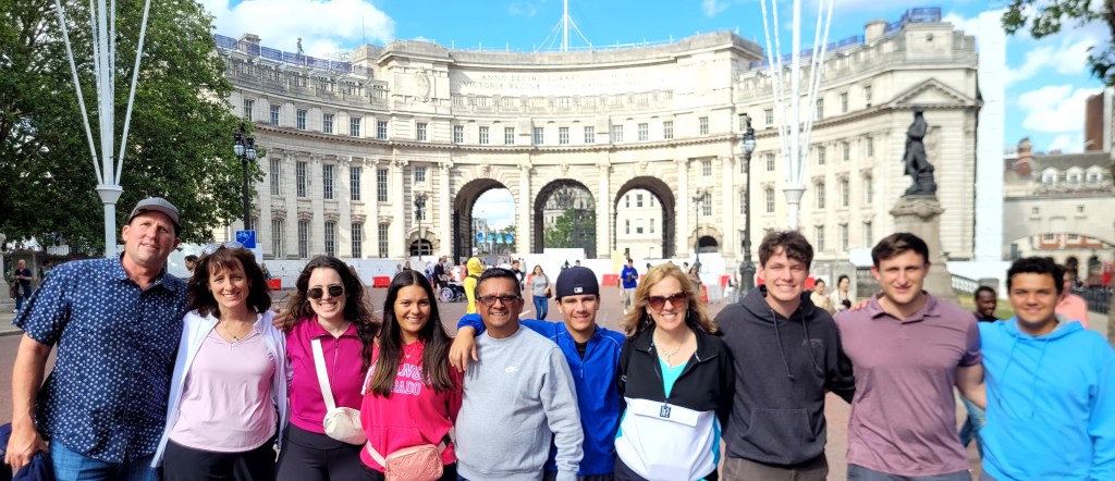 An American family 0f 10 line up in front of Admiralty Arch in London in sunshine, casually dressed for vacation and smiling.